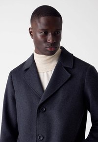 Cappotto di lana grigio scuro con revers, dotato di due grandi bottoni e di un morbido dolcevita crema sottostante. Tessuto liscio e strutturato.