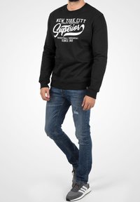 Czarna bluza z białym graficznym napisem, w parze z niebieskimi, dopasowanymi jeansami. Model nosi szare sneakersy z białymi paskami. Gładka tekstura tkaniny.