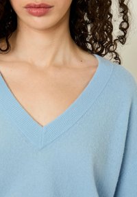 Pull en tricot doux bleu clair avec un col en V. Le tissu a une texture lisse et une coupe décontractée, mettant en valeur le décolleté et les épaules.