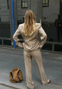 Beige puvun takki tiukasti istuvalla bleiserillä ja leveälahkeisilla housuilla, joissa on ompeluyksityiskohtia. Ruskea olkalaukku aseteltuna maahan.