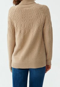 Beżowy sweter z golfem, wykonany z miękkiej tkaniny, z fakturowanym wzorem warkocza na plecach i prążkowanym dołem, zestawiony z niebieskimi dżinsami.