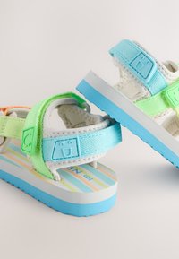 Sandali colorati con suola bianca strutturata e dettagli blu. Cinghie in materiali verde e blu, con sottopiede a righe multicolore.