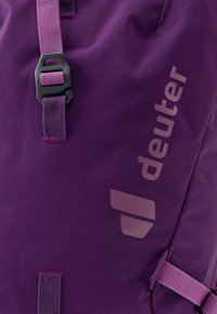 Violeta mugursoma ar teksturētu auduma apdari, aprīkota ar melnu sprādzi un gaišā tonī izdrukātu logotipu. Iekļautas regulējamas siksnas.