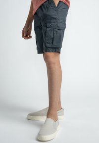 Harmaa cargo-shortsit, joissa on sivutaskut, vyötärövyö ja rento istuvuus, yhdistettynä vaaleanharmaisiin slip-on-kengiin yksinkertaisella taustalla.