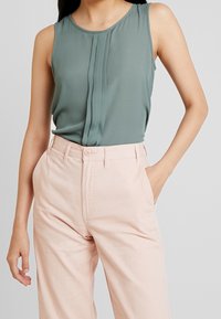 Blusa senza maniche di colore verde scuro con dettagli plissettati sul davanti, abbinata a pantaloni a vita alta di colore rosa chiaro con chiusura a bottone e tasche laterali.
