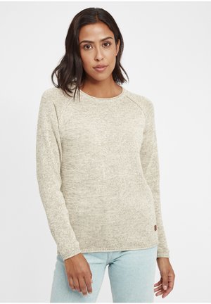 Maglione beige con lavorazione a maglia texture, scollo rotondo e maniche lunghe raglan. Presenta una piccola etichetta logo sul lato inferiore. Abbinato a jeans chiari.