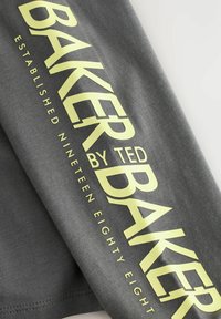 Grauer Stoff mit großen gelben Text, der "BAKER BY TED BAKER" liest, und kleinerem Text "GEGRÜNDET NEUNZEHNHUNDERT ACHTUNDACHTZIG."