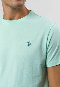 Mátově zelené bavlněné tričko s kulatým výstřihem, s jemným logem v tmavší modré barvě na hrudi a krátkými rukávy. Měkká textura.