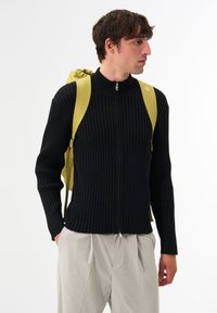 Schwarzer ribbeliger Reißverschluss-Pullover mit hohem Kragen, kombiniert mit einem gelben Rucksack. Das Model trägt hellgraue Hosen. Strukturiertes Material, taillierte Passform.