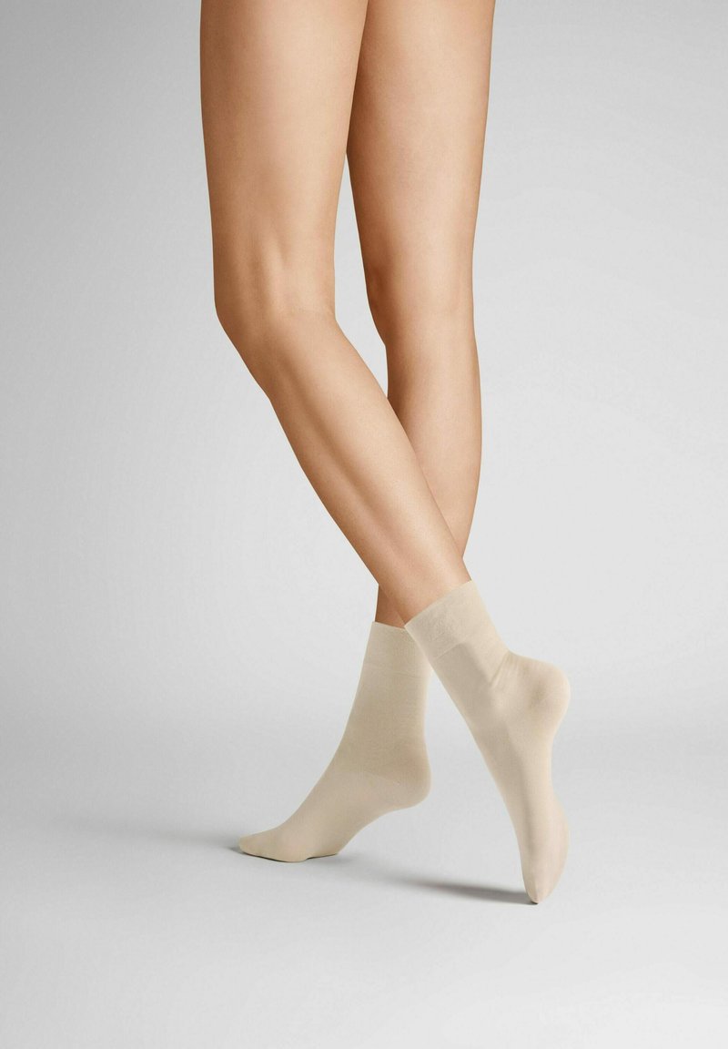 Beige Knöchelsocken aus glattem, dehnbarem Material mit einem gerippten Bündchen, gezeigt auf übereinandergeschlagenen Beinen vor einem neutralen Hintergrund.