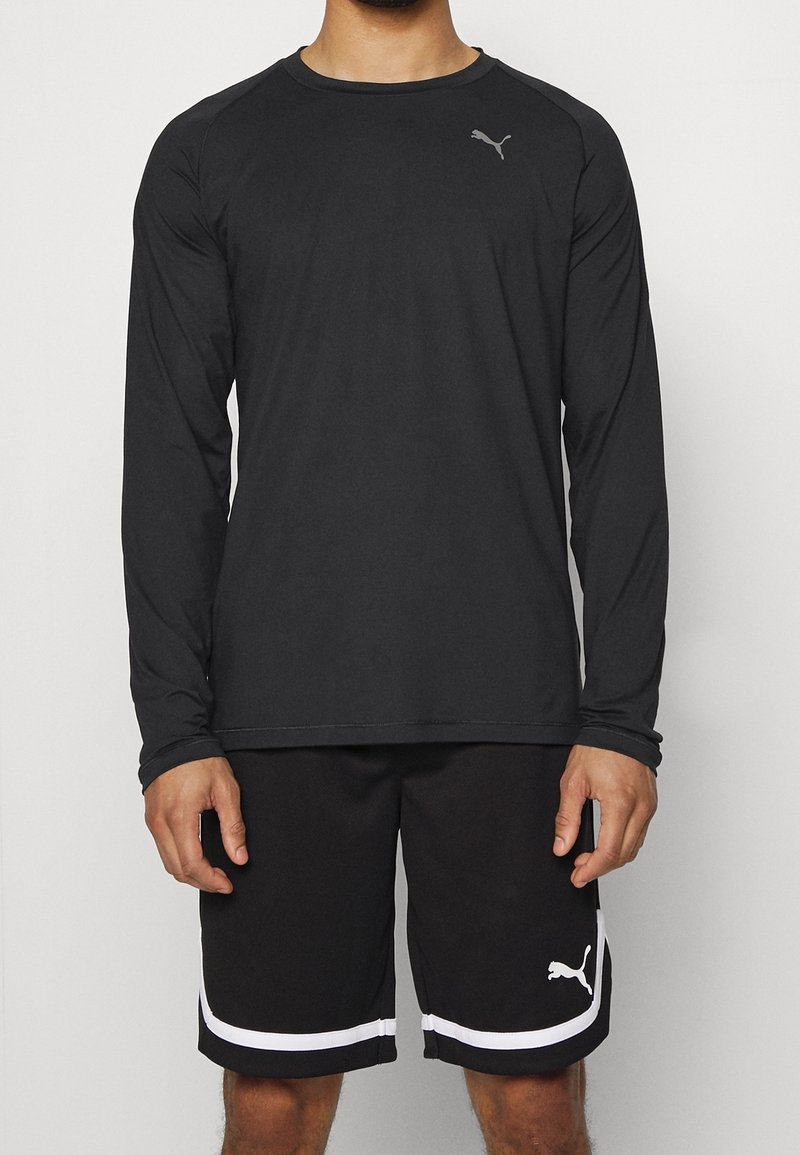 Muž ve černém dlouhém sportovním tričku a černých šortkách s bílými logy Puma a bílými pruhy na šortkách.