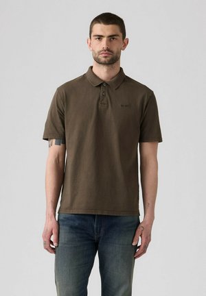 Mann mit kurzen dunklen Haaren, der ein braunes kurzärmeliges Poloshirt und blaue Jeans trägt, steht vor einem schlichten hellen Hintergrund.