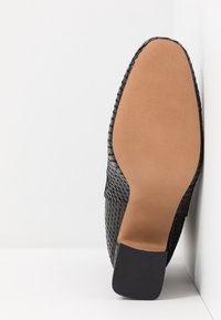 Semelle de chaussure texturée noire avec dessous lisse couleur tan et talon carré noir sur fond de sol et mur blancs.