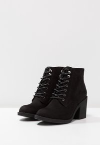 Une paire de bottines en daim noir avec des talons blocs et un laçage sur une surface blanche et un fond gris clair.