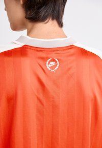 Tricou roșu Nike cu guler alb, având o textură cu nervuri verticale. Prezintă un logo încercuit de lauri pe spate.