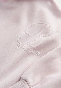 Gaišs rozā sporta krekls ar ribotām piedurknēm un mīkstu tekstūru, uz kura ir izsistais logo "BY TED BAKER" nedaudz tumšākā nokrāsā.