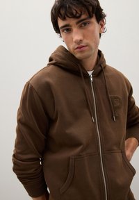 Felpa con zip marrone e cappuccio con coulisse, caratterizzata da una tasca frontale e una lettera "P" testurizzata sul petto. Tessuto liscio.