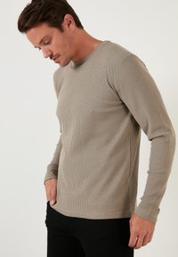 Beige ribbed long-sleeve shirt with a round neckline and fitted design, worn over black pants. Texture visible, highlighting the fabric's details.