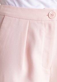 Detailní záběr na světle růžové texturované kalhoty s záhybem a odpovídajícím růžovým knoflíkem na pase.