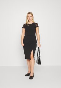Schwarzes, figurbetontes Kleid mit kurzen Spitzenärmeln und seitlichem Schlitz. Das Model hält eine schwarze Handtasche und trägt schwarze High Heels. Einfaches, elegantes Design.