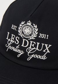 Black cap featuring white embroidered text reading "LES DEUX Sporting Goods" and a decorative emblem with laurel leaves and a crown.