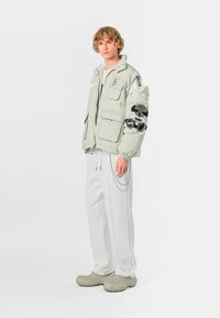 Chaqueta acolchada verde claro con múltiples bolsillos y detalles bordados en negro, combinada con pantalones blancos y calzado texturizado gris.