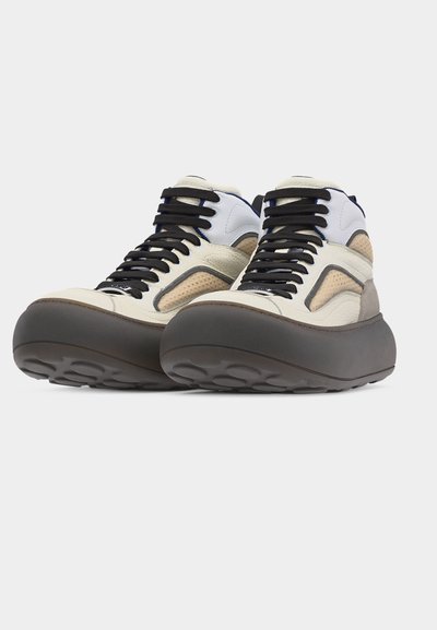 Vysoké tenisky v béžovej a bielej farbe s čiernymi šnúrkami a hrubými masívnymi tmavohnedými podrážkami, zobrazené z predného uhla.