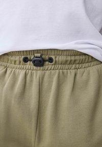 Khaki joggers elastsega vööga, musta reguleeritava nööriga ja pehme tekstuuriga, asetatud valge särke alla.