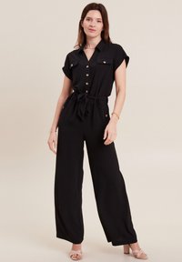 Zwarte jumpsuit van lichtgewicht stof, met korte mouwen, een knoopsluiting aan de voorkant, twee borstzakken en een strikceintuur in de taille.