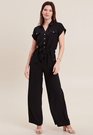 Combinaison noire en tissu léger, avec des manches courtes, une fermeture à boutons sur le devant, deux poches poitrine et une ceinture à nouer à la taille.