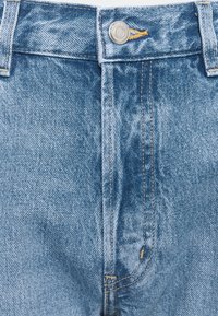 Detailní pohled na přední stranu modrých džínových kalhot se stříbrným knoflíkem a viditelnými švy na pásku a oblasti zipu.