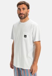 Bílý tričko s krátkým rukávem vyrobené z bavlny, s přední kapsou a černou nálepkou. Model má na sobě pruhované šortky pro kontrast.