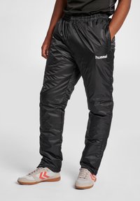 Schwarze, glänzende, leichte Trainingshosen mit einem elastischen Bund und Taschen. Mit weißem Logo und Seitennähten mit subtiler Textur.
