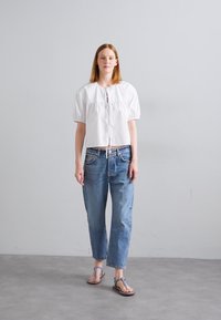 Bluză albă scurtă cu mâneci umflate și un nod în față, combinată cu blugi din denim de culoare albastră deschisă și sandale brune cu întreținere. Texturi netede.