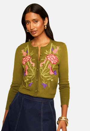 Vrouw draagt een olijfgroen vest met roze en paarse bloemrijke borduursels, gouden creolen, gouden armbanden en een donkerblauwe rok.