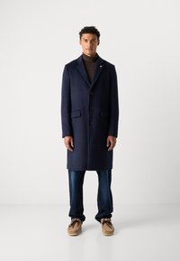 Abrigo de marina hecho de lana con solapa, presenta tres botones y dos bolsillos laterales, combinado con jeans oscuros y zapatos beige.