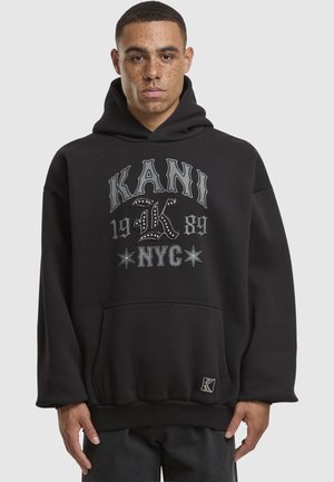 Schwarzer Kapuzenpullover mit Fronttasche, der in grauer Schrift "KANI 1989 NYC" und dekorativen Nieten gestaltet ist. Lockerer Schnitt, gerippte Bündchen.