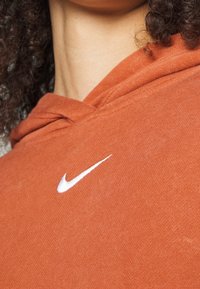 Oranžová mikina s kapucí vyrobená z bavlny, s bílým logem Nike na hrudi a stahovac�í kapucí. Detailní pohled na látku.