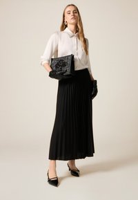 Valkoinen nappipaita, musta liehuhame, mustat käsineet ja kiiltävän nahkaiset korot. Kädessä kuvioitu musta clutch-laukku.