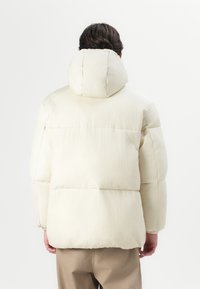 Bílá prošívaná bunda s kapucí, s prošívanou texturou a objemnou siluetou. Hladká látka s viditelnými stehmi.