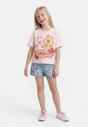 Smilende pige iført en pink T-shirt med to tegneserie-capybaraer, der drikker juice, og denimshorts med jordbærprint, står på hvid baggrund.