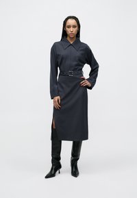 Tmavozelené košeľové šaty s dlhými rukávmi, veľkým golierom a bočnými rozparkami; v páse stiahnuté opaskom so striebornou obdĺžnikovou prackou.