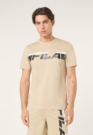 Młody mężczyzna w beżowej koszulce marki FILA oraz dopasowanych szortach z czarno-białymi paskami z logo, stojący na jednolitym tle.