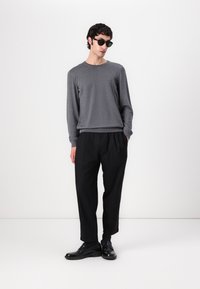 Pull en gris à manches longues avec un tricot texturé, associé à un pantalon noir ample et des chaussures en cuir noires. Le mannequin porte des lunettes de soleil sombres.