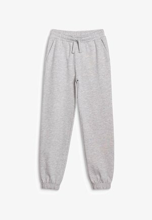 Fille souriante debout, portant un sweat à capuche gris clair, un pantalon de survêtement assorti, des baskets blanches et un bandeau blanc sur un fond uni.