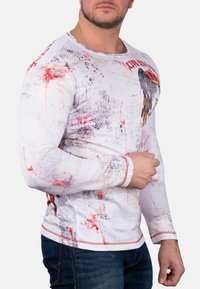 Langarm-Shirt in Weiß mit rot- und graufarbigen Farbspritzern. Verfügt über einen runden Ausschnitt und kontrastierende rote Nähte an den Bündchen.