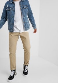 Person wearing light brown pants, white t-shirt, blue denim jacket, and black-and-white high-top sneakers standing against a plain white background.