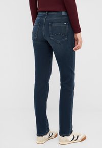 Sötétkék farmernadrág, slim fit szabással, két hátsó zsebbel és diszkrét varrási részletekkel. Bézs tornacipővel párosítva.