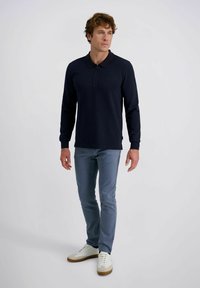 Mann steht da und trägt ein navyblaues Langarm-Polohemd, hellblaue Jeans und weiße Sneaker vor einem schlichten weißen Hintergrund.