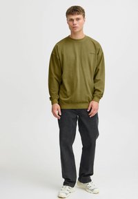Oliwkowy sweatshirt z okrągłym dekoltem, z prążkowanymi mankietami i dołem. Noszony z ciemnymi dżinsami i białymi trampkami. Prosty design, bez wzorów.